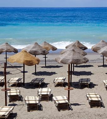 Spiaggia con ombrelloni di paglia, lettini vuoti, mare azzurro e onde delicate.