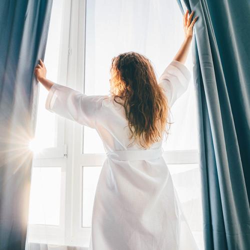 Donna apre le tende, abbracciando la luce del sole mattutino con serenità e speranza.